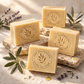 Four bars of The Australian Olive Oil Soap on a stone surface with olive branches and lavender flowers. THEAOOS