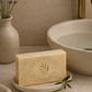 Bar of soap on a stone dish with a vase of lavender and a sink in the background.. The AUSTRALIAN OLIVE OIL SOAP