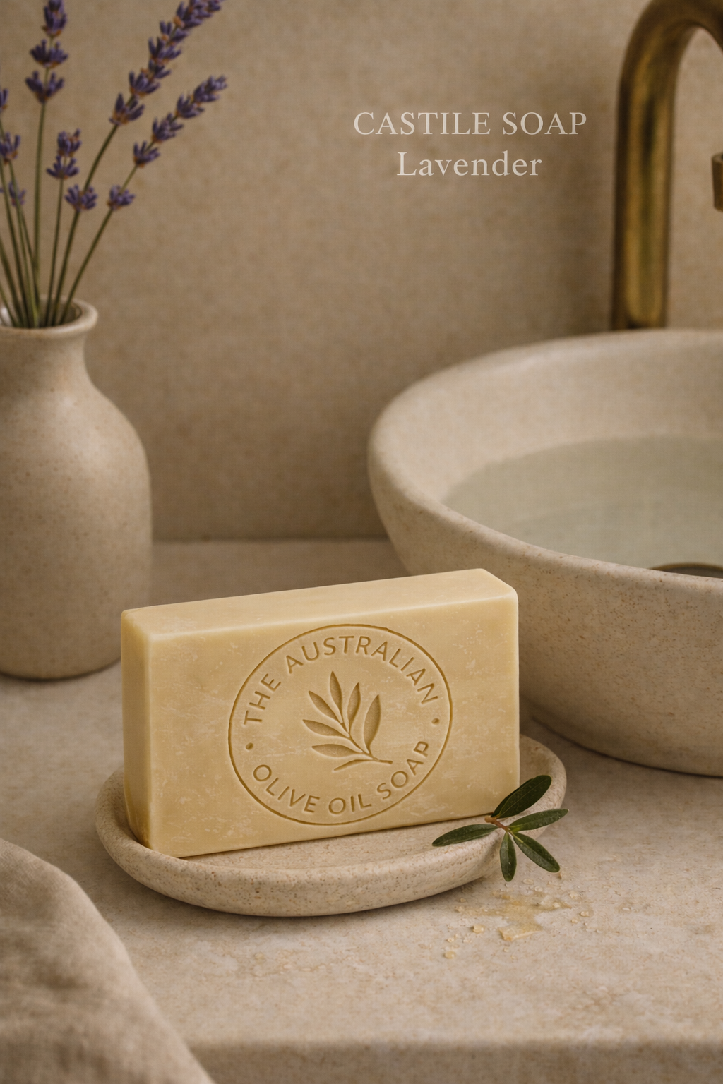 Bar of soap on a stone dish with a vase of lavender and a sink in the background.. The AUSTRALIAN OLIVE OIL SOAP