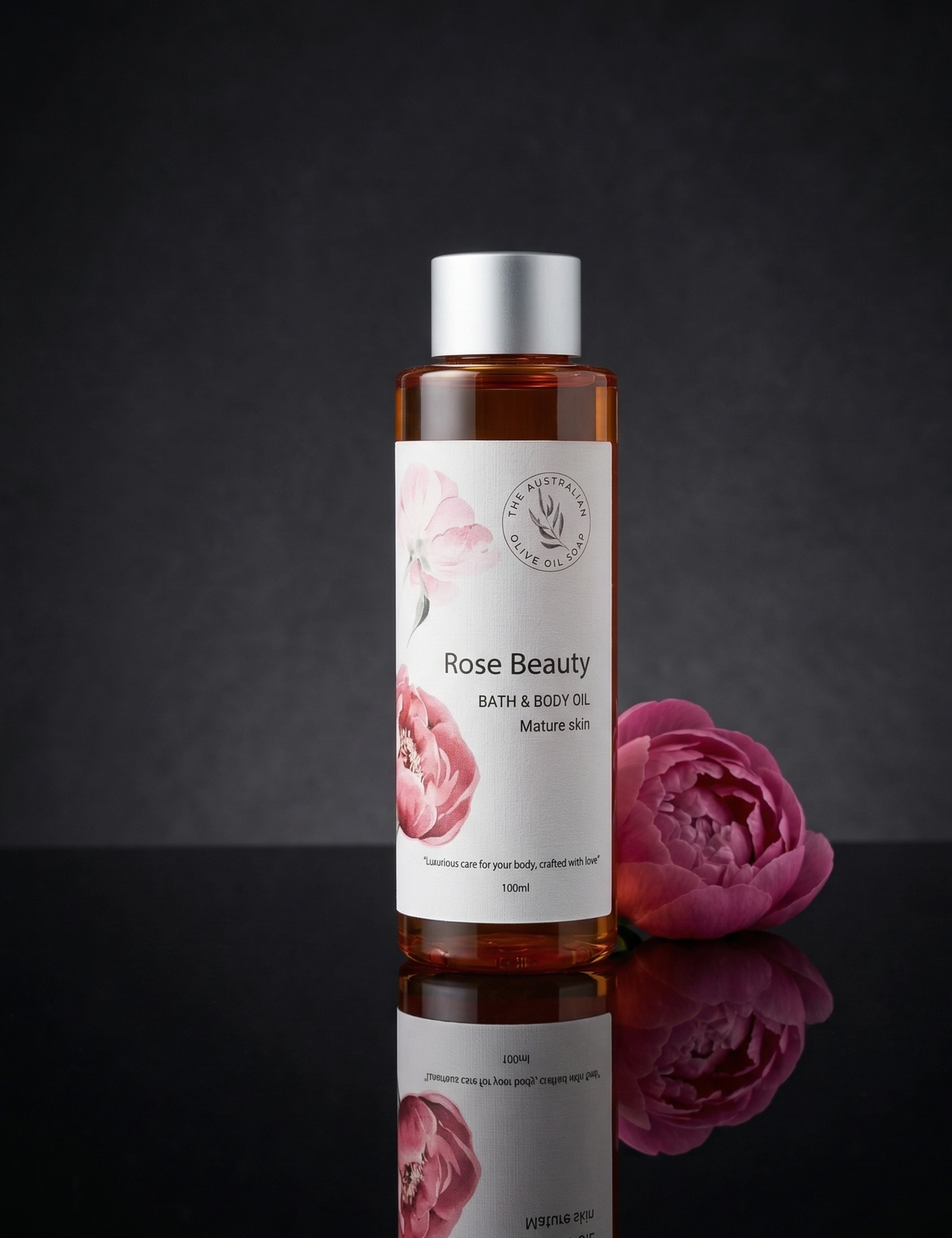 Bottle of 'Rose Beauty' bath and body oil with a pink flower on a dark background- The Australian Olive Oil Soap