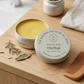 Olive oil balm in a tin with a wooden comb and herbs on a wooden surface