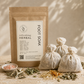 A kraft paper pouch labeled 'Refreshing Herbal Foot Soak' stands on a stone surface beside three small drawstring burlap bags filled with herbs, along with loose dried herbs and a small dish of pink salt. The scene is styled with warm natural shadows and no person or animal present.
