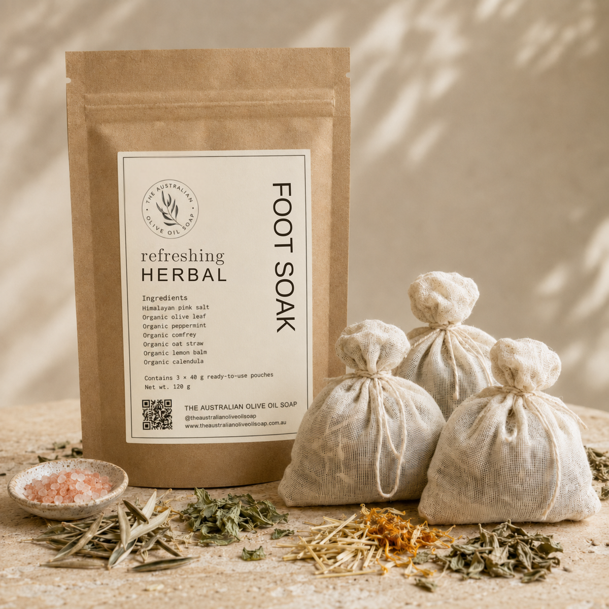 A kraft paper pouch labeled 'Refreshing Herbal Foot Soak' stands on a stone surface beside three small drawstring burlap bags filled with herbs, along with loose dried herbs and a small dish of pink salt. The scene is styled with warm natural shadows and no person or animal present.