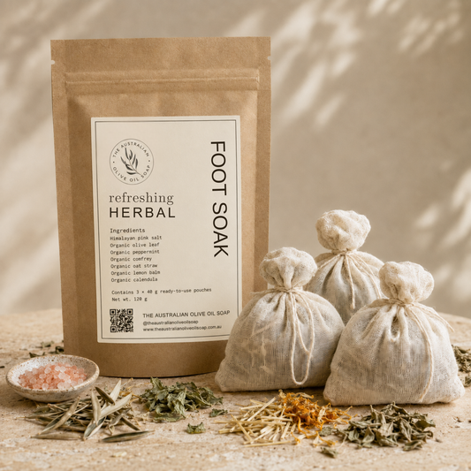 A kraft paper pouch labeled 'Refreshing Herbal Foot Soak' stands on a stone surface beside three small drawstring burlap bags filled with herbs, along with loose dried herbs and a small dish of pink salt. The scene is styled with warm natural shadows and no person or animal present.
